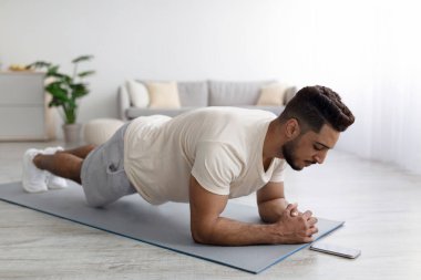 Ev içi güç egzersizi. Genç bir Arap cep telefonundaki video dersini takip ederek evde dirsek tahtası yapıyor.