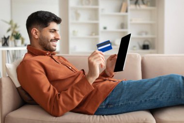 Tablet ve kredi kartı kullanan gülümseyen adam koltukta oturuyor.