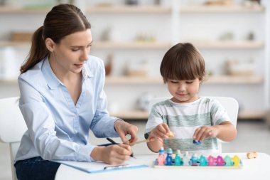 Uzman ofisinde renkli ahşap oyunlar oynayan sevimli küçük çocuk, not alan profesyonel kadın terapist, boş alan.