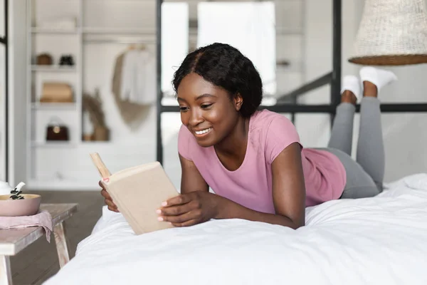 Черная женщина читает книгу, лежащую на животе в спальне — стоковое фото