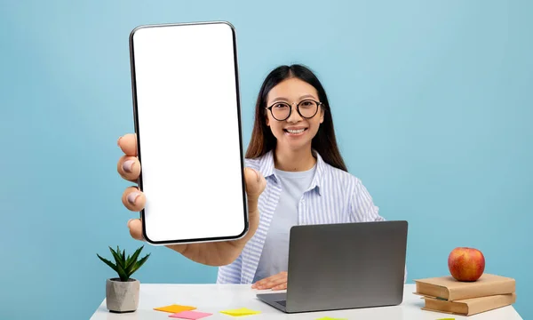 Felice studentessa asiatica dimostrando grande smartphone con schermo bianco, seduta sul posto di lavoro su sfondo blu — Foto Stock