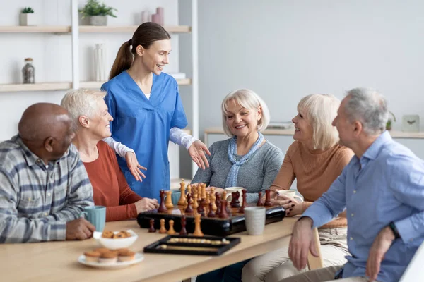 Enfermeira amigável falando com pessoas idosas jogando jogos de mesa — Fotografia de Stock