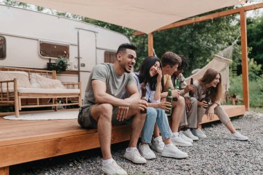 Mutlu çokuluslu arkadaşlar bira içiyor, sohbet ediyor, gülüyor, karavanın yanında oturuyor, kamp yaparken eğleniyorlar.