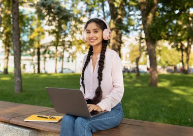 Parkta dizüstü bilgisayarı ve kulaklığı olan mutlu Hintli kadın, bankta oturuyor ve dışarıda kameraya gülümsüyor.