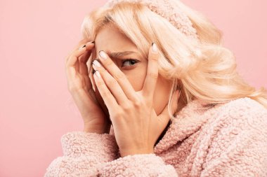 Şok olmuş genç bayan korkuyla yüzünü kapatıyor, pembe stüdyo arka planında duruyor.