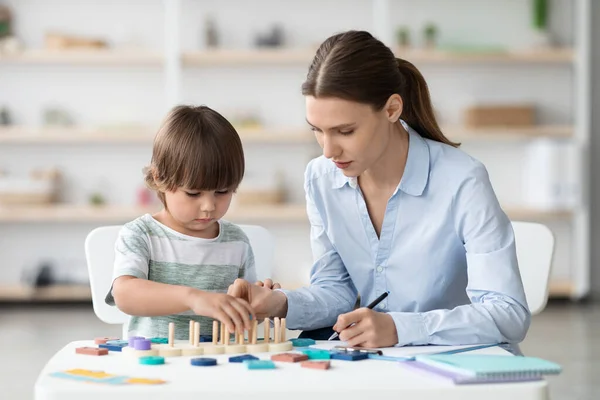 Schattig jongetje speelt logisch spel bij vakkabinet, vrouwelijke psycholoog maakt notities op kantoor, lege ruimte — Stockfoto