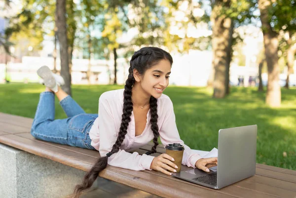 Descansa en el campus. Chica estudiante universitaria india feliz relajarse al aire libre con café y computadora portátil, acostado en el banco en el parque — Foto de Stock