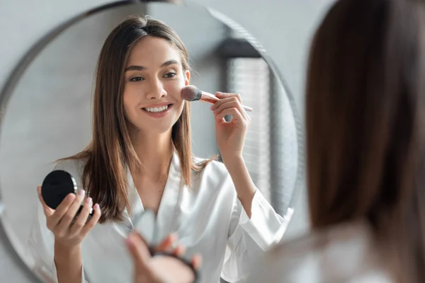 Привлекательная молодая женщина делает ежедневный макияж, стоя возле зеркала в ванной комнате — стоковое фото