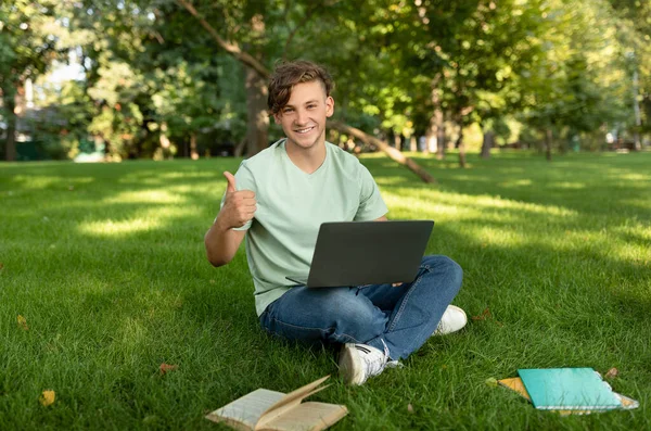 Счастливый студент-мужчина жестикулирует, одобряет образовательную онлайн-программу, сидит с ноутбуком в парке, показывает большой палец вверх — стоковое фото