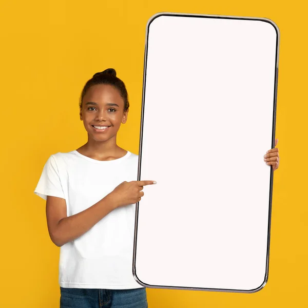 可愛いです黒十代の女の子指しています巨大スマートフォンとともに空の画面上のオレンジの背景,モックアップのためのアプリデザイン — ストック写真