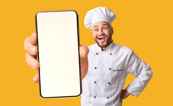 Happy šéfkuchař ukazuje smartphone s bílou prázdnou obrazovkou, doporučuje catering služby nebo dodávání potravin app — Stock fotografie