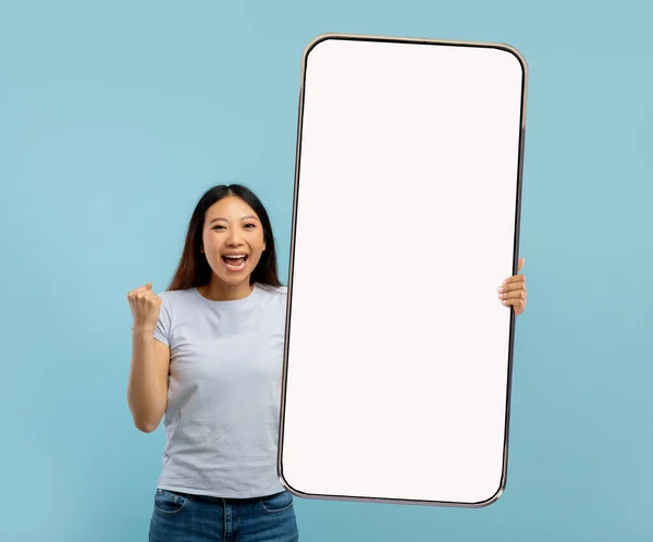 Jeune femme asiatique excitée tenant un gros téléphone portable avec écran vide, montrant le geste OUI, offrant une maquette pour l'annonce — Photo