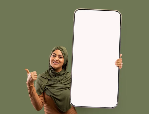年轻漂亮的阿拉伯女人，头戴头巾，手里拿着大手机，屏幕空空如也，露出大拇指，摆出一副笑脸 — 图库照片
