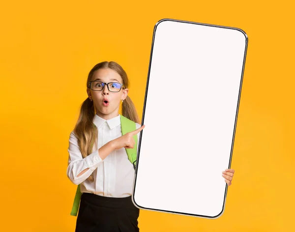 Escolar impactada con mochila apuntando a un gran teléfono celular con pantalla vacía, que ofrece maqueta para la aplicación de estudio en línea — Foto de Stock