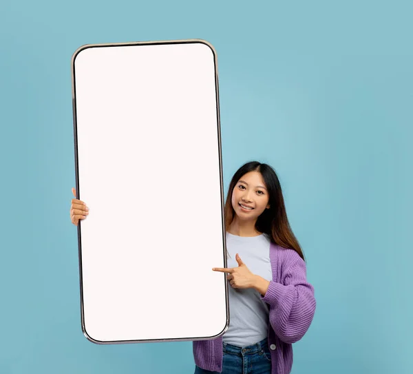 Sonriente mujer asiática joven apuntando a un gran teléfono celular con pantalla vacía sobre fondo de estudio azul. Burla. —  Fotos de Stock