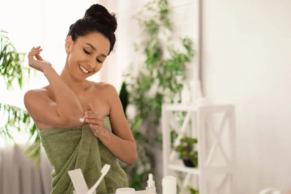 Ładna milenijna kobieta w ręczniku nakładająca krem uspokajający na łokcie przed lustrem w domu. Leczenie suchej skóry — Zdjęcie stockowe