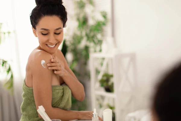 Beautiful millennial woman applying cream on her shoulder, taking care of skin after bath near mirror at home, copy space