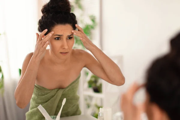 Concerned millennial female looking at her reflection in mirror, checking first wrinkles on forehead at bathroom, free space — Stock Photo, Image