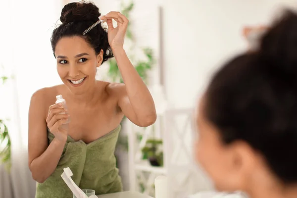 Mujer joven y alegre en toalla aplicando suero o aceite natural en su cabello cerca del espejo en casa, espacio para copiar — Foto de Stock