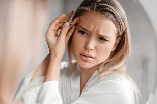 Eliminación del exceso de vello de la cara. Mujer joven en ropa de dormir arrancando cejas en el interior del baño, primer plano —  Fotos de Stock