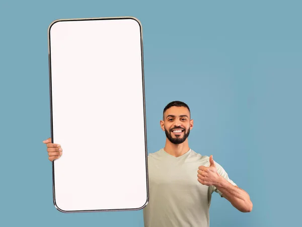 Lächelnder junger arabischer Mann demonstriert riesiges Smartphone mit Attrappe, zeigt Daumen hoch, wirbt für Anwendung oder Website — Stockfoto