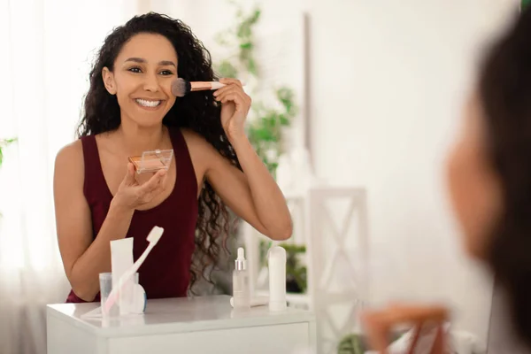 Hermosa mujer de pie frente al espejo, sosteniendo cepillo y aplicando maquillaje, polvo facial o bronceador en el baño —  Fotos de Stock