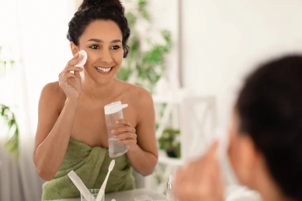 Söt ung brunett kvinna bär handduk efter bad, uppfriskande hennes hud med lotion eller tonic nära spegel hemma — Stockfoto
