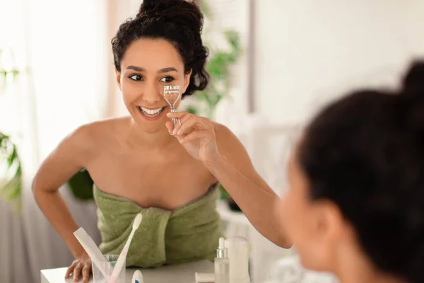 Mulher milenar muito sorridente na toalha usando curler cílios perto do espelho em casa, espaço de cópia — Fotografia de Stock