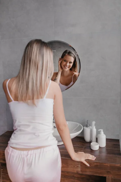 Jovencita en pijama mirando al espejo y tocándose la cara, aplicando cosméticos hidratantes en su piel y sonriendo —  Fotos de Stock