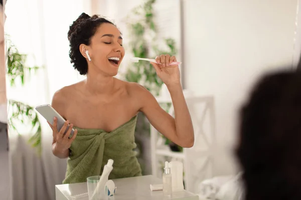 Mujer joven feliz en toalla usando teléfono inteligente y auriculares, cantando en el cepillo de dientes, divirtiéndose en casa — Foto de Stock