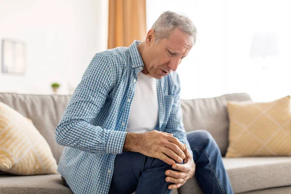 Dojrzały mężczyzna z bólem kolana siedzi na kanapie w domu — Zdjęcie stockowe