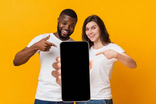 Gunakan aplikasi baru ini. Happy multirasial pasangan menunjuk pada smartphone dengan layar kosong, mockup untuk desain — Stok Foto