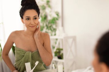 Havlu giymiş neşeli genç bir kadın banyo ve duştan sonra yüzüne dokunuyor ve mükemmel cildine hayran. Aynanın yanında duruyor.