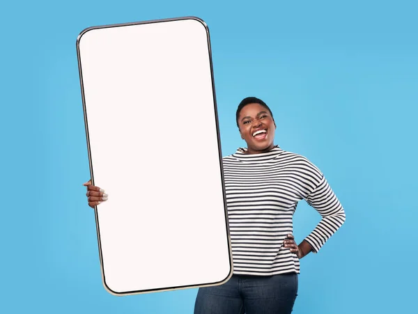 身材魁梧的年轻黑人女士在蓝色工作室的背景上展示了一个空白屏幕的大型智能手机，这是对网络广告的嘲弄 — 图库照片
