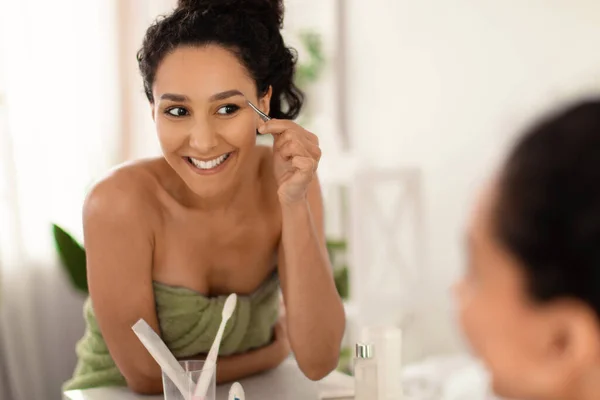 Sorrindo jovem mulher usando toalha, corrigindo sobrancelhas com pinça perto do espelho em casa, espaço livre — Fotografia de Stock