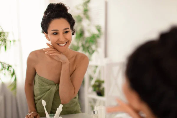 Preciosa mujer milenaria con una perfecta piel suave tocando la cara, mimándose después de la ducha o el baño, mirándose en el espejo — Foto de Stock