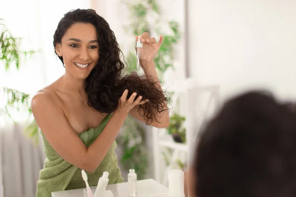 Encantadora jovem morena aplicando soro no cabelo seco danificado perto do espelho em casa, espaço em branco — Fotografia de Stock