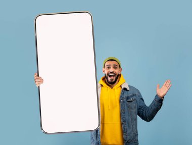 Heyecanlı bin yıllık Arap adam mavi stüdyo arka planında dev akıllı telefon ekranında yeni mobil uygulama için modellik teklif ediyor