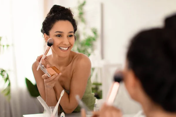 Χαμογελώντας νεαρή γυναίκα εφαρμογή μακιγιάζ μπροστά από τον καθρέφτη, βάζοντας σε σκόνη προσώπου, blusher ή bronzer στο σπίτι — Φωτογραφία Αρχείου