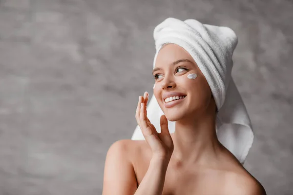 Női bőr koncepció. Lady alkalmazása hidratáló krém a szem alatt, áll a fürdőszobában, félrenéz és mosolyog — Stock Fotó