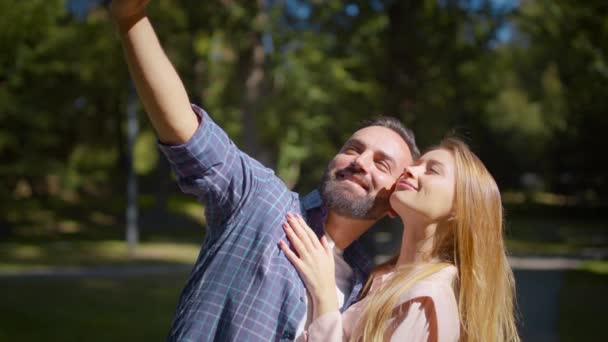 Щаслива пара в любові робить селфі на смартфоні, позує разом у зеленому парку під час літньої дати — стокове відео