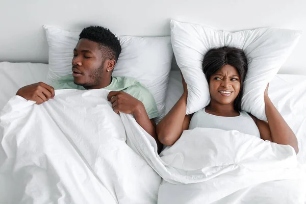 Mujer afroamericana joven infeliz cubriéndose las orejas con almohada, sufriendo de ronquidos de marido dormido —  Fotos de Stock