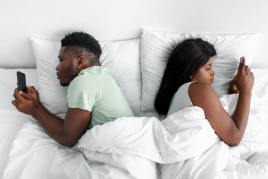 Ciddi Afro-Amerikalı kadın ve erkek sırt üstü beyaz yatağa uzanıp yatak odasında akıllı telefonlara yazı yazıyorlar.