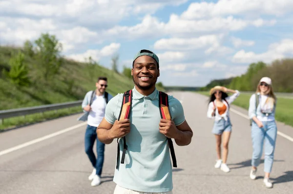 Szczęśliwy czarny facet z wielonarodowymi przyjaciółmi spacerujących wzdłuż autostrady, zabawy podczas podróży autostopem lato — Zdjęcie stockowe