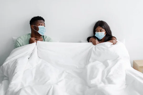 Triste infeliz joven negro enfermo esposa y marido en protección máscaras en social distanciamiento en cama bajo blanco manta —  Fotos de Stock