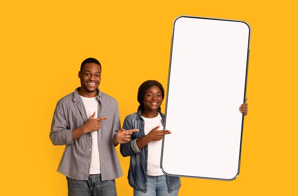 Épouses afro-américaines excitées pointant vers un grand smirtphone avec écran blanc vierge, recommandant votre annonce, maquette — Photo