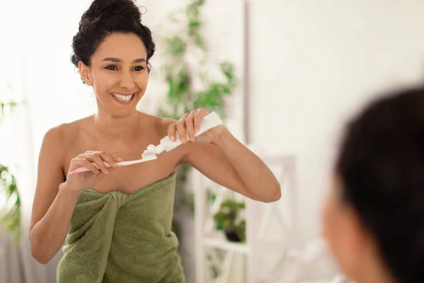 Ελκυστική νεαρή γυναίκα με υπέροχο χαμόγελο φορώντας πετσέτα μετά το ντους, εφαρμόζοντας οδοντόκρεμα σε πινέλο κοντά στον καθρέφτη στο σπίτι — Φωτογραφία Αρχείου