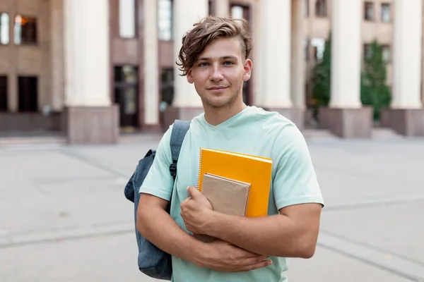Πορτραίφ ενός χαρούμενου φοιτητή με σακίδιο πλάτης και τετράδια που ποζάρουν πάνω από πανεπιστημιακό υπόβαθρο — Φωτογραφία Αρχείου