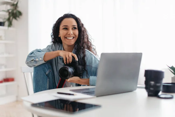 Mujer fotógrafa feliz sosteniendo la cámara fotográfica usando computadora portátil sentada en interiores —  Fotos de Stock