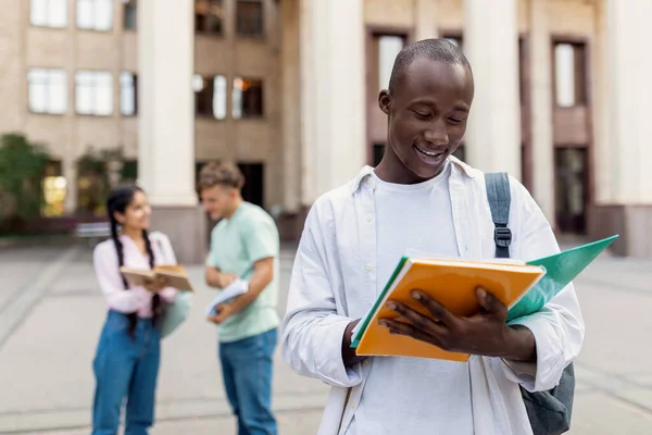 Preto estudante universitário cara segurando notebook e leitura, de pé ao ar livre com seus colegas de classe em segundo plano — Fotografia de Stock